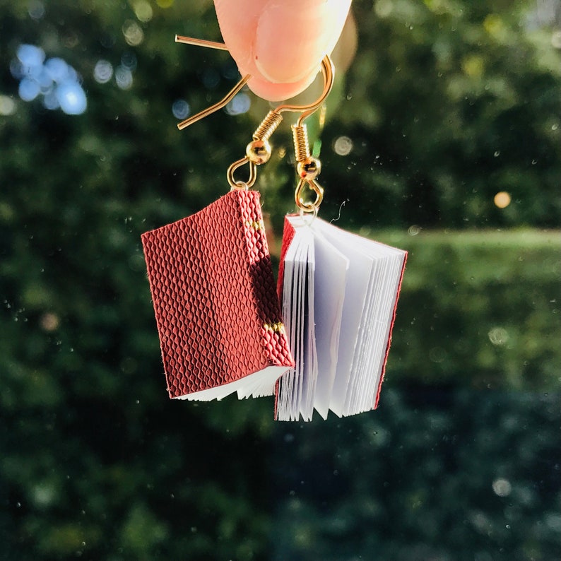Op de afbeelding: Oorbellen in de vorm van een boek. Elke oorbel heeft een miniatuurboekje met een getextureerde, bordeauxrode kaft en witte pagina's. De boeken hangen aan goudkleurige oorhaken, wat een uniek literair accessoire cre&euml;ert.