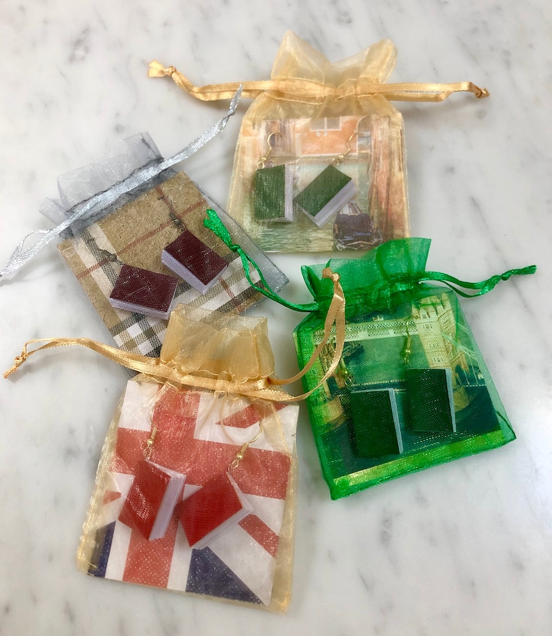 Op de afbeelding: Vier kleine organza zakjes, elk met een paar oorbellen in de vorm van een boek. De zakjes zijn in zilver, goud en groen, met de oorbellen in rood, groen en bruin. De zakjes hebben verschillende bedrukte achtergronden.