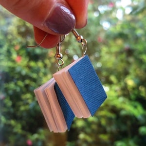 Op de afbeelding: Een paar unieke oorbellen in de vorm van miniatuurboeken. Elke oorbel heeft een blauwe, getextureerde kaft en witte pagina's, opgehangen aan zilverkleurige oorhaakjes. De achtergrond is een vage groene omgeving.