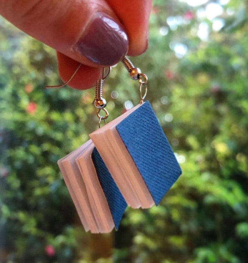 Op de afbeelding: Een paar unieke oorbellen in de vorm van miniatuurboeken. Elke oorbel heeft een blauwe, getextureerde kaft en witte pagina's, opgehangen aan zilverkleurige oorhaakjes. De achtergrond is een vage groene omgeving.