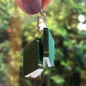 Op de afbeelding: Hangende oorbellen met miniatuur groene boekbedels met gouden accenten. De kleine boeken hebben witte pagina's en zijn opgehangen aan zilverkleurige oorhaken. De achtergrond is een wazig beeld van groen gebladerte.