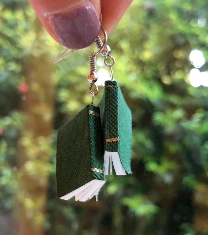 Op de afbeelding: Hangende oorbellen met miniatuur groene boekbedels met gouden accenten. De kleine boeken hebben witte pagina's en zijn opgehangen aan zilverkleurige oorhaken. De achtergrond is een wazig beeld van groen gebladerte.