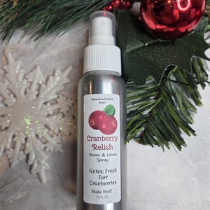 May include: A silver spray bottle labeled "Cranberry Relish Room & Linen Spray" with a cranberry illustration. The label lists "Fresh Tart Cranberries" as the scent notes. The bottle is 2.5 fl oz and is surrounded by Christmas decorations.