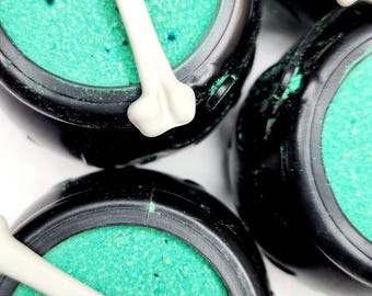 May include: A collection of black plastic cauldron-shaped containers filled with a vibrant green textured substance. Each cauldron is topped with a single off-white bone-shaped accent piece resting across the top of the green contents.