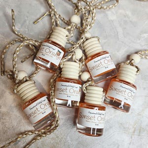 May include: Six small glass bottles of Sunset Orange fragrance with wooden caps and white labels. Each bottle is suspended by a gold and white rope with a wooden bead. The bottles contain an orange-colored liquid and are labeled with the scent name and volume.