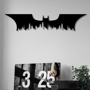 Op de afbeelding: Zwarte metalen Batman-silhouet wanddecoratie met een stadsbeeldontwerp. De decoratie is gemonteerd op een witte muur boven een bureau met een computer, koptelefoon en een bruine leren bureaustoel.