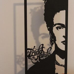 May include: Black and white paper cut-out portrait of Frida Kahlo with flowers in her hair. The portrait is framed in black paper.