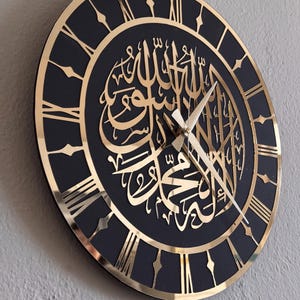 May include: A black and gold wall clock with Roman numeral markings and Arabic calligraphy in gold. The clock has a black background and gold accents.