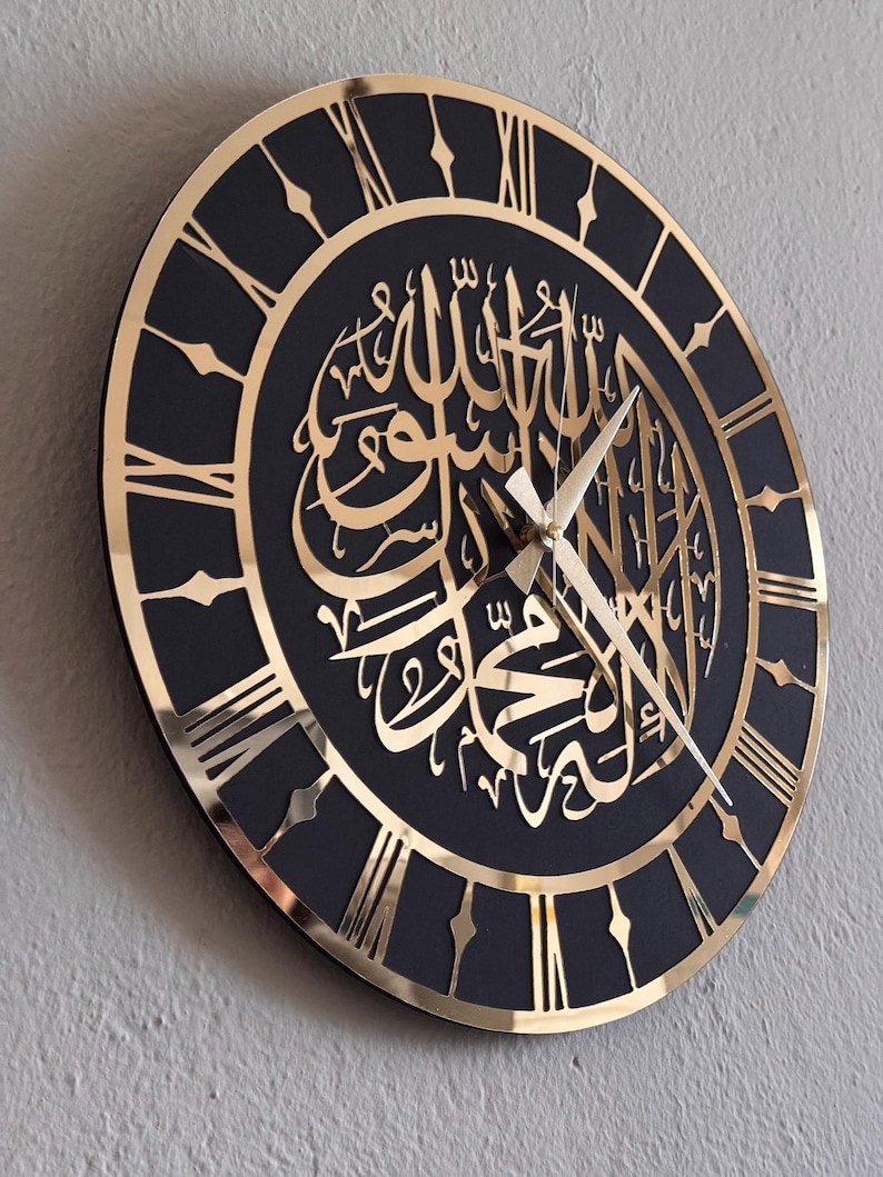 May include: A black and gold wall clock with Roman numeral markings and Arabic calligraphy in gold. The clock has a black background and gold accents.