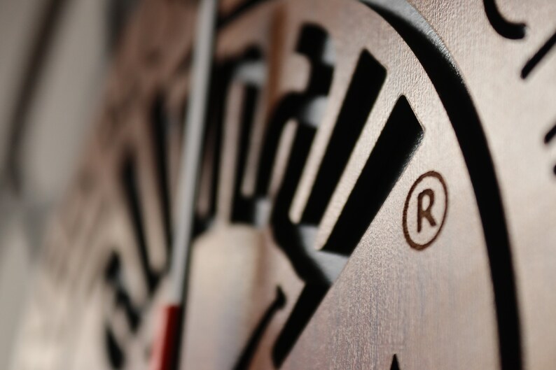 May include: Close-up of a wooden clock face with black lettering and a registered trademark symbol. The clock face is made of wood with a light brown finish.