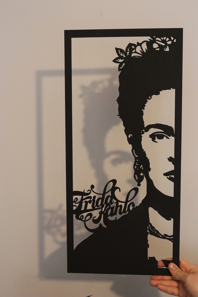May include: Black and white paper cut-out portrait of Frida Kahlo with flowers in her hair. The portrait is framed in black paper.