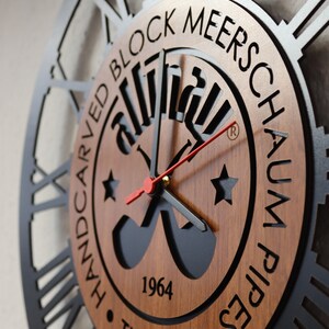 May include: A black and brown wooden wall clock with Roman numerals and a black metal frame. The clock face features a logo with the text "Handcarved Block Meerschaum Pipes 1964 Turkey".