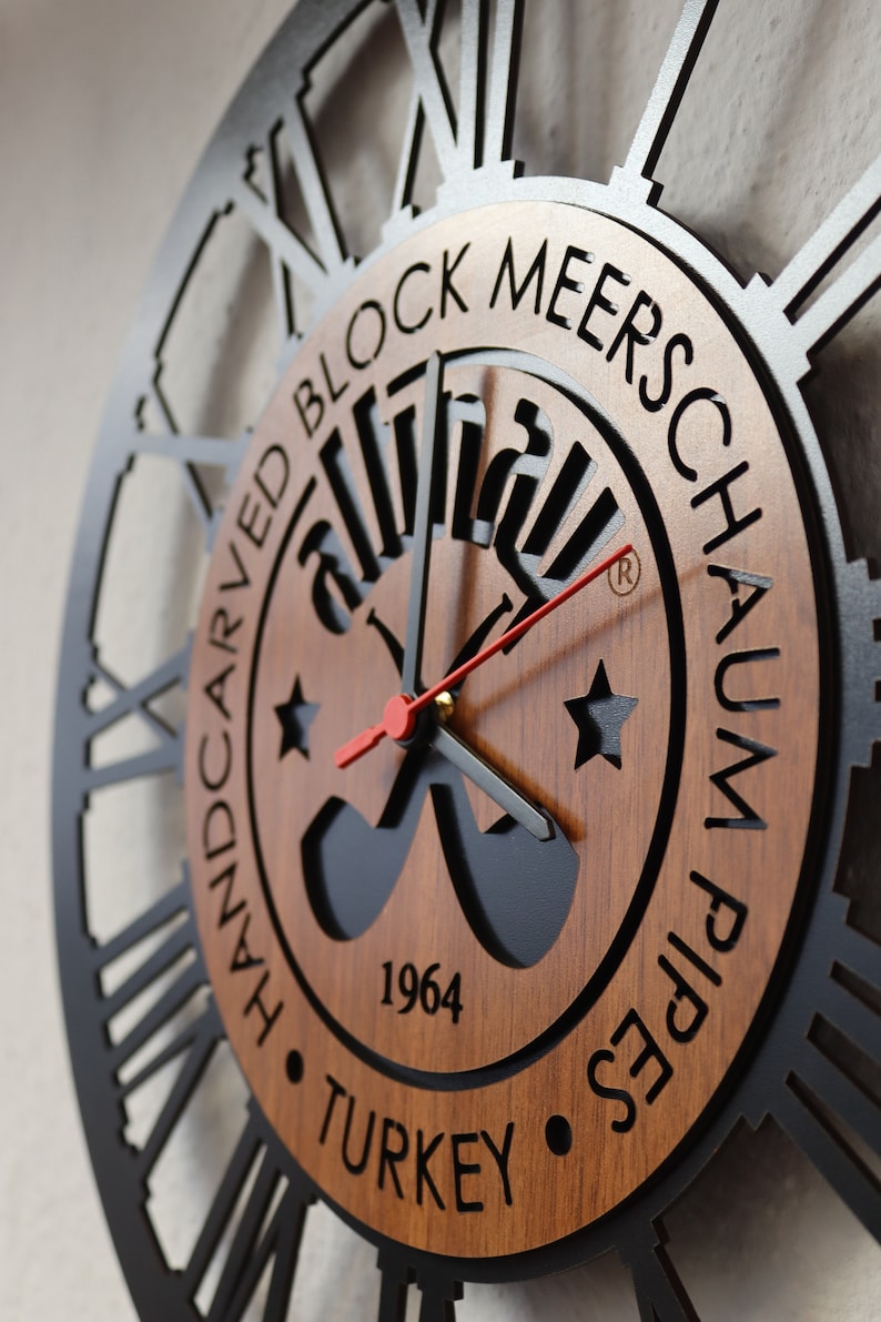 May include: A black and brown wooden wall clock with Roman numerals and a black metal frame. The clock face features a logo with the text "Handcarved Block Meerschaum Pipes 1964 Turkey".