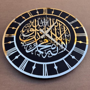 May include: A black and gold clock with Roman numerals and Arabic calligraphy in gold. The clock has a black background and a gold rim.