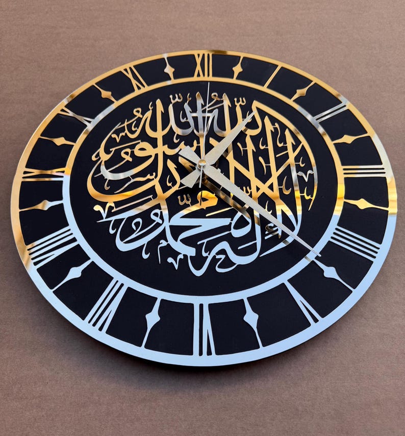 May include: A black and gold clock with Roman numerals and Arabic calligraphy in gold. The clock has a black background and a gold rim.