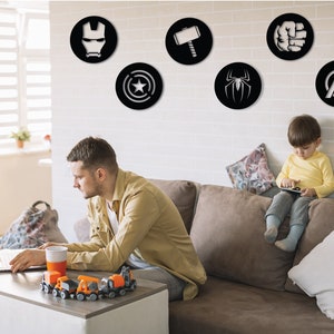 Puede incluir: Una sala de estar con una pared de ladrillo blanco decorada con seis calcoman&iacute;as circulares de pared negras y blancas con logotipos de superh&eacute;roes. Un hombre est&aacute; sentado en un sof&aacute; marr&oacute;n usando una computadora port&aacute;til. Un ni&ntilde;o peque&ntilde;o est&aacute; sentado en el sof&aacute; jugando con una tableta.