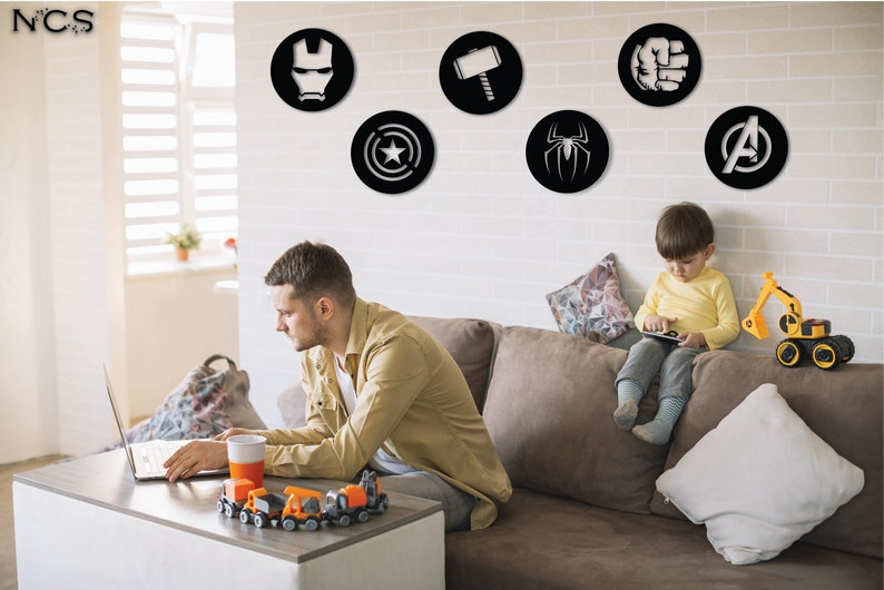 Puede incluir: Una sala de estar con una pared de ladrillo blanco decorada con seis calcoman&iacute;as circulares de pared negras y blancas con logotipos de superh&eacute;roes. Un hombre est&aacute; sentado en un sof&aacute; marr&oacute;n usando una computadora port&aacute;til. Un ni&ntilde;o peque&ntilde;o est&aacute; sentado en el sof&aacute; jugando con una tableta.