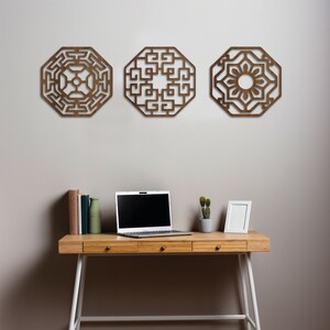 May include: Three wooden wall decorations in the shape of octagons with intricate geometric designs. The pieces are hanging on a light grey wall above a wooden desk with a laptop, books, a vase, and a small potted plant.