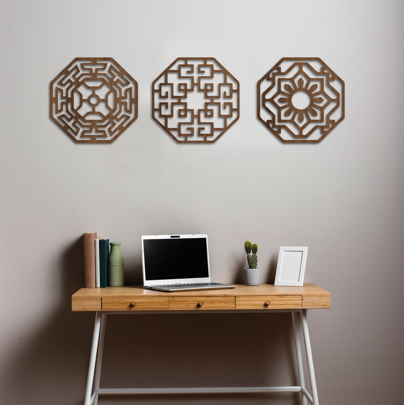 May include: Three wooden wall decorations in the shape of octagons with intricate geometric designs. The pieces are hanging on a light grey wall above a wooden desk with a laptop, books, a vase, and a small potted plant.