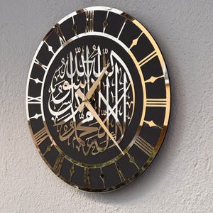 May include: A black and gold clock with Roman numerals and Arabic calligraphy. The clock has a black background and gold accents. The calligraphy is in Arabic and is a religious inscription.