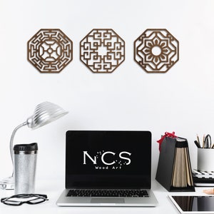 May include: Three brown wooden wall decorations with intricate geometric designs. The designs are in a traditional Asian style. The decor is hanging on a white wall. A laptop with the text "NCS Wood Art" is on a desk in the foreground.
