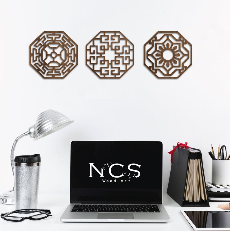 May include: Three brown wooden wall decorations with intricate geometric designs. The designs are in a traditional Asian style. The decor is hanging on a white wall. A laptop with the text "NCS Wood Art" is on a desk in the foreground.