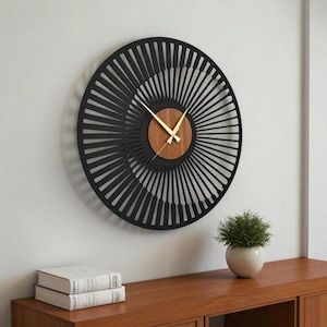 May include: A modern, round wall clock with a black, radial design. The clock features a wooden center and gold-colored hands. The clock is mounted on a white wall, with a wooden cabinet and a small potted plant visible below.
