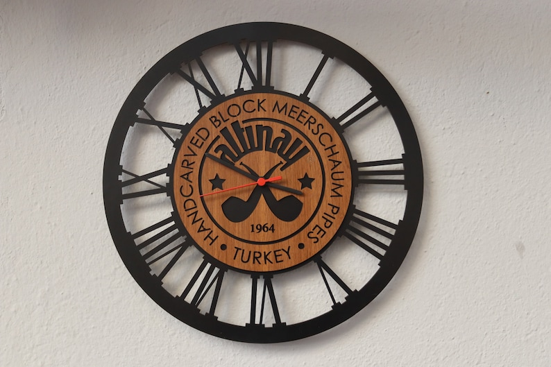 May include: Black and brown wooden wall clock with Roman numeral markings. The clock face features a logo with the text "Handcarved Block Meerschaum Pipes" and "1964 Turkey".