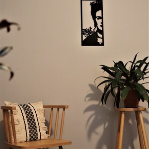 May include: Black metal wall art featuring a silhouette of Frida Kahlo with the text "Frida Kahlo" in a stylized font. The art is hanging on a white wall with a wooden chair and a potted plant in the foreground.