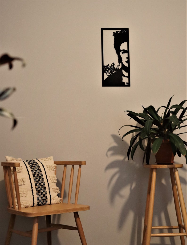 May include: Black metal wall art featuring a silhouette of Frida Kahlo with the text "Frida Kahlo" in a stylized font. The art is hanging on a white wall with a wooden chair and a potted plant in the foreground.