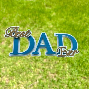 May include: A decorative cake topper featuring the text Best DAD Ever. The word DAD is in large, bold blue letters with a white outline. The words Best and Ever are in a smaller, cursive gold font with a white outline, attached to a clear stick.
