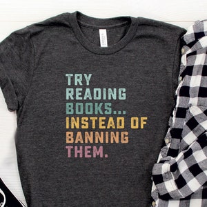 May include: A dark grey t-shirt with the text "TRY READING BOOKS...INSTEAD OF BANNING THEM." printed in colorful letters. The shirt is laid flat on a white wooden surface with a pair of black sunglasses, a black and white plaid shirt, and two black canvas shoes.