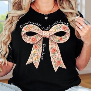 May include: Black t-shirt featuring a large bow design with floral accents in pink, yellow, and orange. The bow has a woven texture. The text "Mom she is clothed with strength dignity" and "Proverbs 31:25" are printed on the shirt.