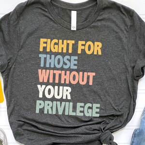 May include: Dark heather gray t-shirt with the text "Fight for those without your privilege" in colorful block letters. The shirt is shown on a model wearing blue jeans.