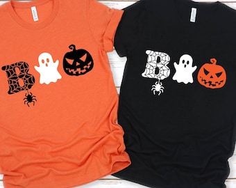 Könnte beinhalten: Zwei T-Shirts mit Halloween-Motiv. Eines ist orange mit dem Wort "BOO", das mit einer Spinne, einem Geist und einem Kürbis ausgeschrieben ist. Das andere ist schwarz mit dem gleichen Design in Weiß.