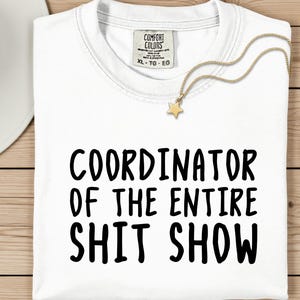 A white t-shirt with the text "COORDINATOR OF THE ENTIRE SHIT SHOW" printed on it, along with a gold star charm. The t-shirt is laid out on a wooden surface, and there is a white hat and a white plate nearby.
