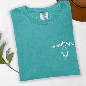 A turquoise t-shirt with a mountain and penguin graphic printed on it, along with a brown hat and a green plant.