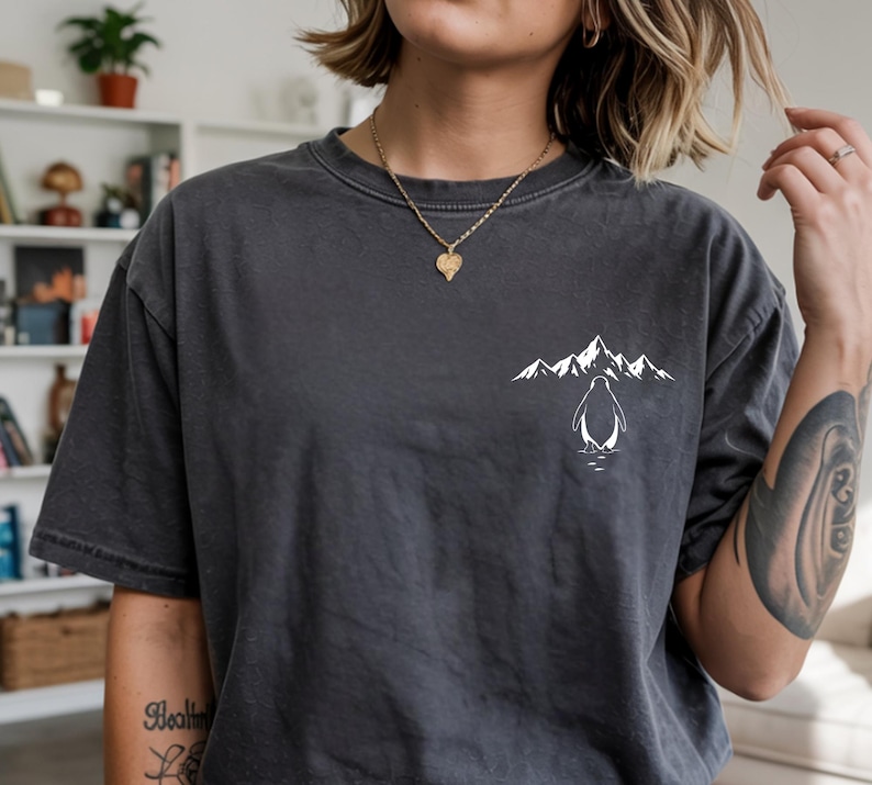 A person wearing a gray t-shirt with a graphic design of a penguin and mountains printed on it.