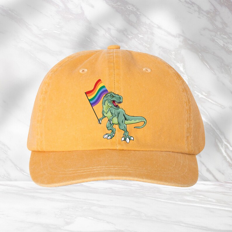 Puede incluir: Una gorra de b&eacute;isbol amarilla con un dinosaurio verde sosteniendo una bandera arco&iacute;ris. El dinosaurio est&aacute; mirando hacia el lado derecho de la gorra.
