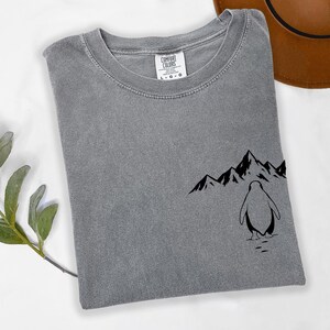 a gray t-shirt with a graphic design of a penguin and mountains on the front, along with a brown hat placed next to it.
