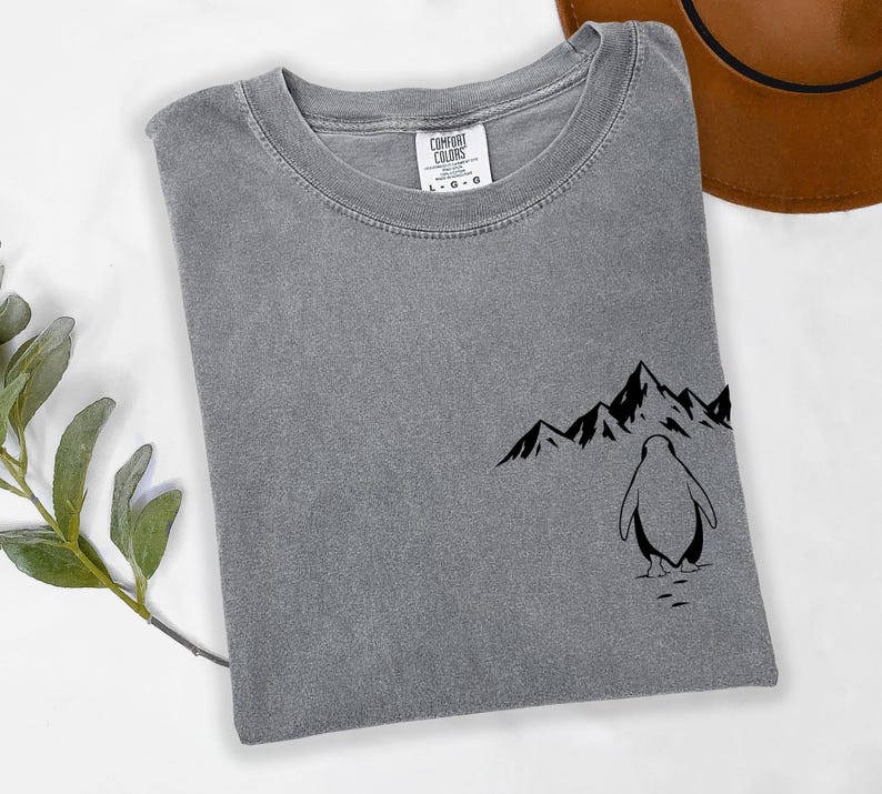 a gray t-shirt with a graphic design of a penguin and mountains on the front, along with a brown hat placed next to it.