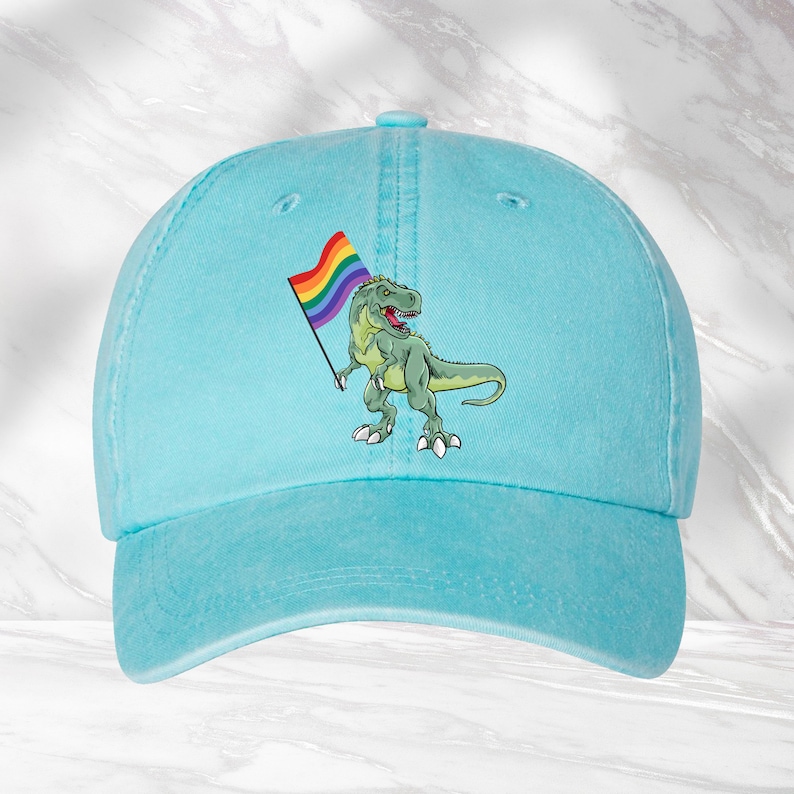 Puede incluir: Una gorra de b&eacute;isbol azul claro con un dinosaurio verde sosteniendo una bandera arco&iacute;ris del orgullo.
