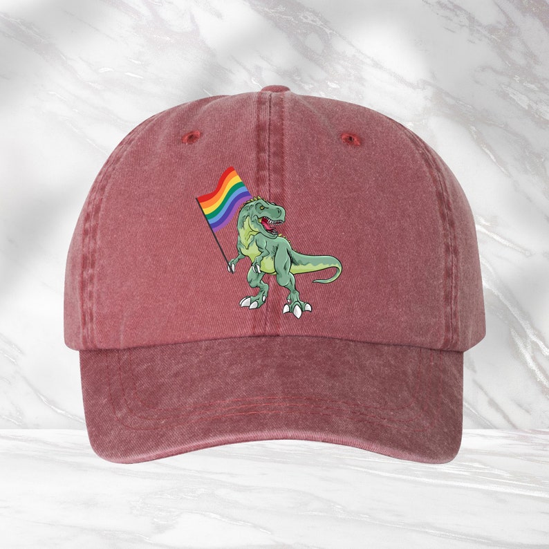 Puede incluir: Una gorra de b&eacute;isbol roja con un dinosaurio verde sosteniendo una bandera arco&iacute;ris del orgullo.