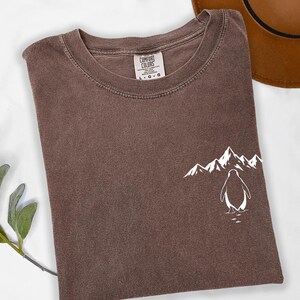 a brown t-shirt with a white graphic of a penguin and mountains on the front, along with a brown hat placed next to it.