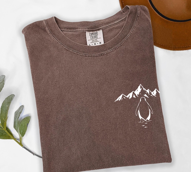 a brown t-shirt with a white graphic of a penguin and mountains on the front, along with a brown hat placed next to it.