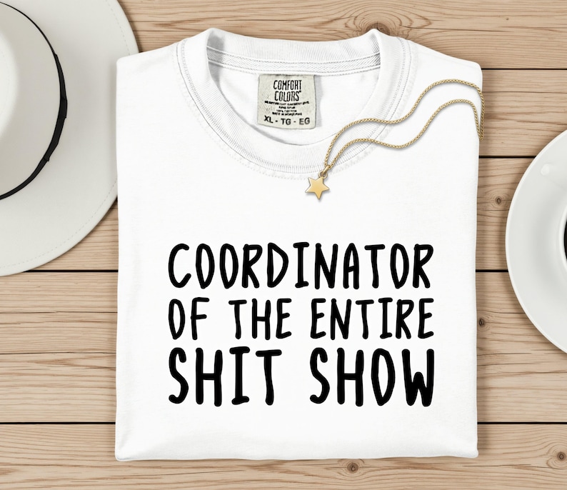 A white t-shirt with the text "COORDINATOR OF THE ENTIRE SHIT SHOW" printed on it, along with a gold star charm. The t-shirt is laid out on a wooden surface, and there is a white hat and a white plate nearby.