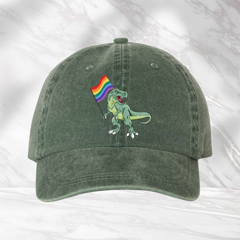 Puede incluir: Una gorra de b&eacute;isbol verde con un dinosaurio de dibujos animados sosteniendo una bandera arco&iacute;ris del orgullo. El dinosaurio es verde con un contorno blanco. La bandera es arco&iacute;ris con un contorno blanco.