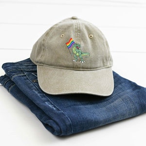 Puede incluir: Una gorra de b&eacute;isbol de color caqui con una imagen bordada de un dinosaurio verde sosteniendo una bandera arco&iacute;ris.
