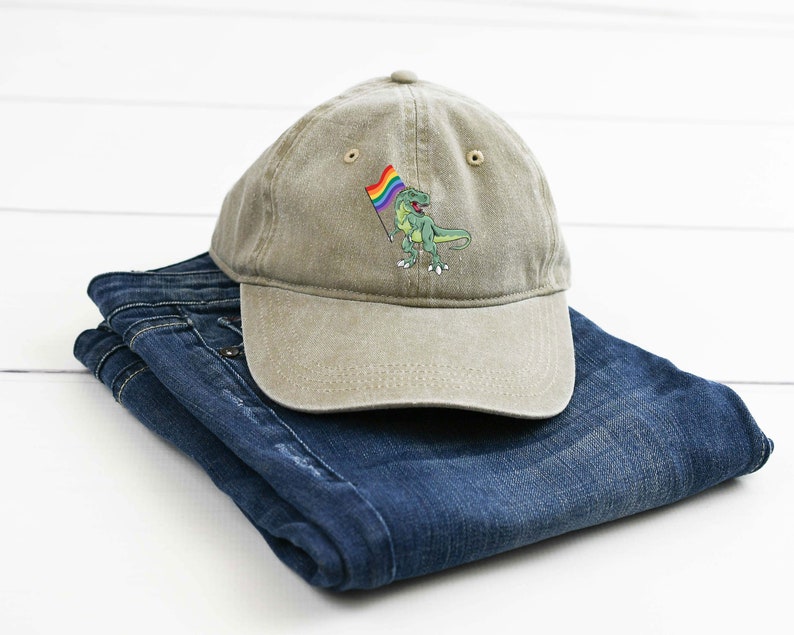 Puede incluir: Una gorra de b&eacute;isbol de color caqui con una imagen bordada de un dinosaurio verde sosteniendo una bandera arco&iacute;ris.