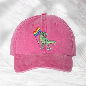 Puede incluir: Gorra de b&eacute;isbol rosa con un dinosaurio verde sosteniendo una bandera arco&iacute;ris.